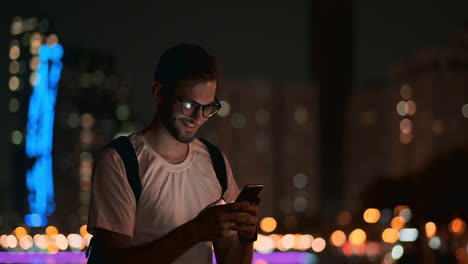A-male-traveler-with-a-backpack-with-glasses-on-the-background-of-the-night-city-calls-a-taxi-through-a-mobile-phone.-Holds-a-smartphone-and-touches-the-screen.-See-photos-and-write-messages-in-social-networks